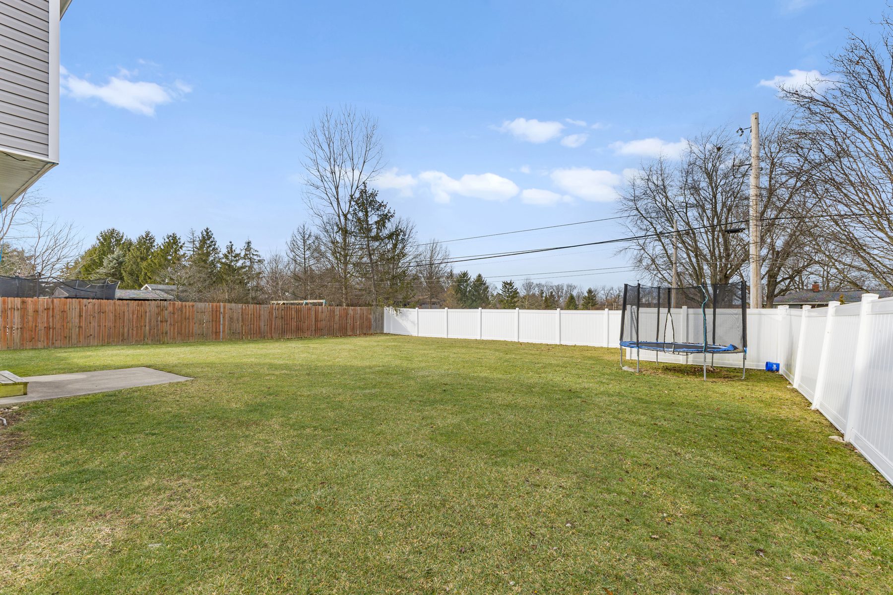 Fenced back yard with mown grass, a trampoline at the far corner and vinyl and wood fencing along the boundary