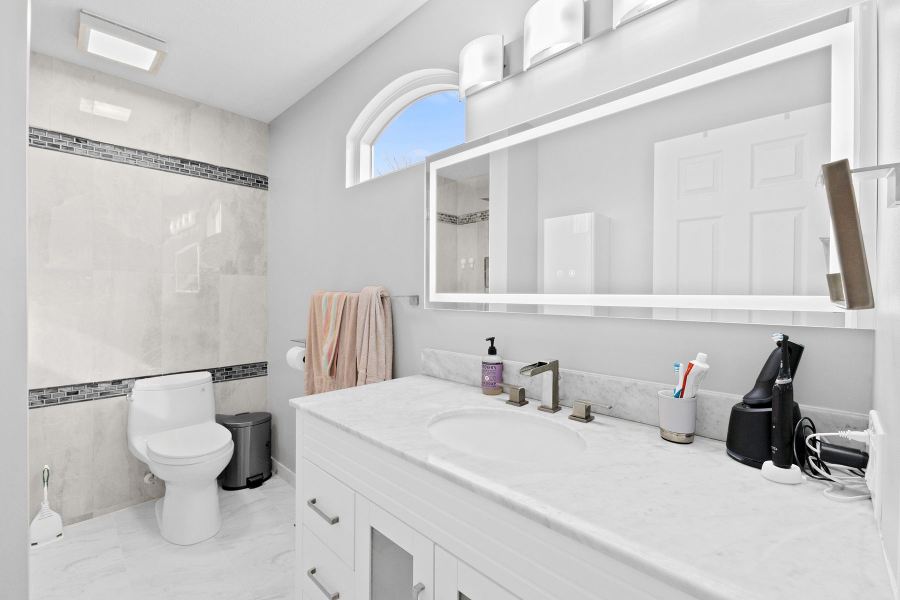 Bathroom with a long white vanity, stone-look counter, wide mirror and tiled walls