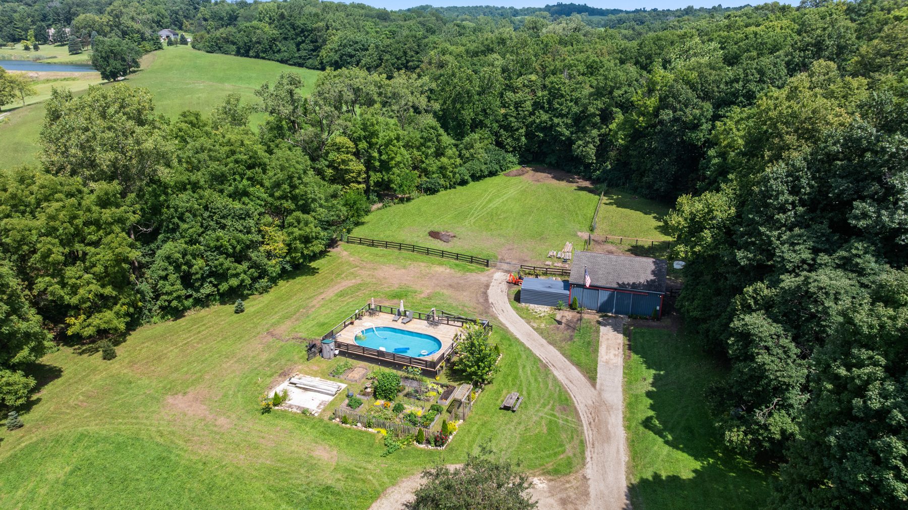 Aerial view from further back showing the house, the above-ground pool and deck, gardens and drive set inside mown lawn and surrounding woods