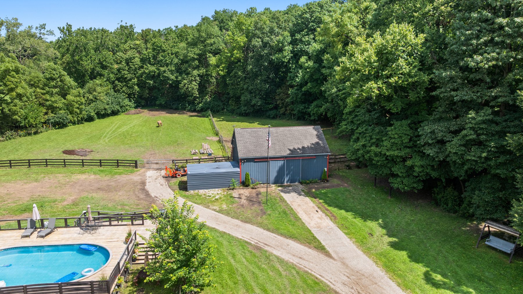 Aerial view low over the pool deck, the fenced paddock beside it and the outbuilding at the end of the drive