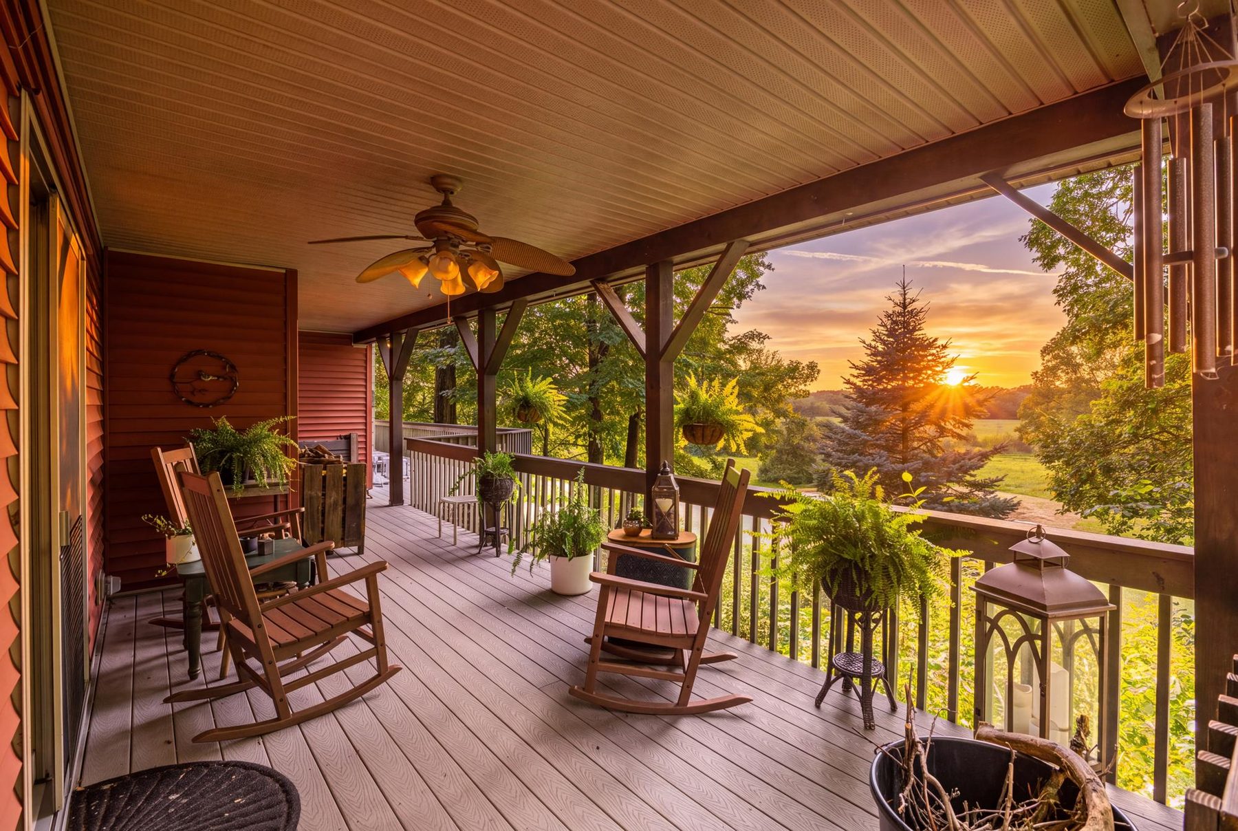 Covered wrap-around porch at sunset, with rocking chairs, hanging ferns and the sun going down behind the tree line