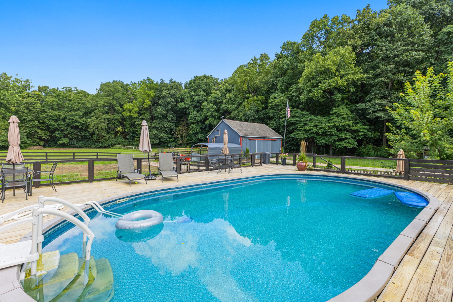 The above-ground pool from the external steps down off its wraparound deck, with loungers, umbrellas and the outbuilding and fenced paddock beyond