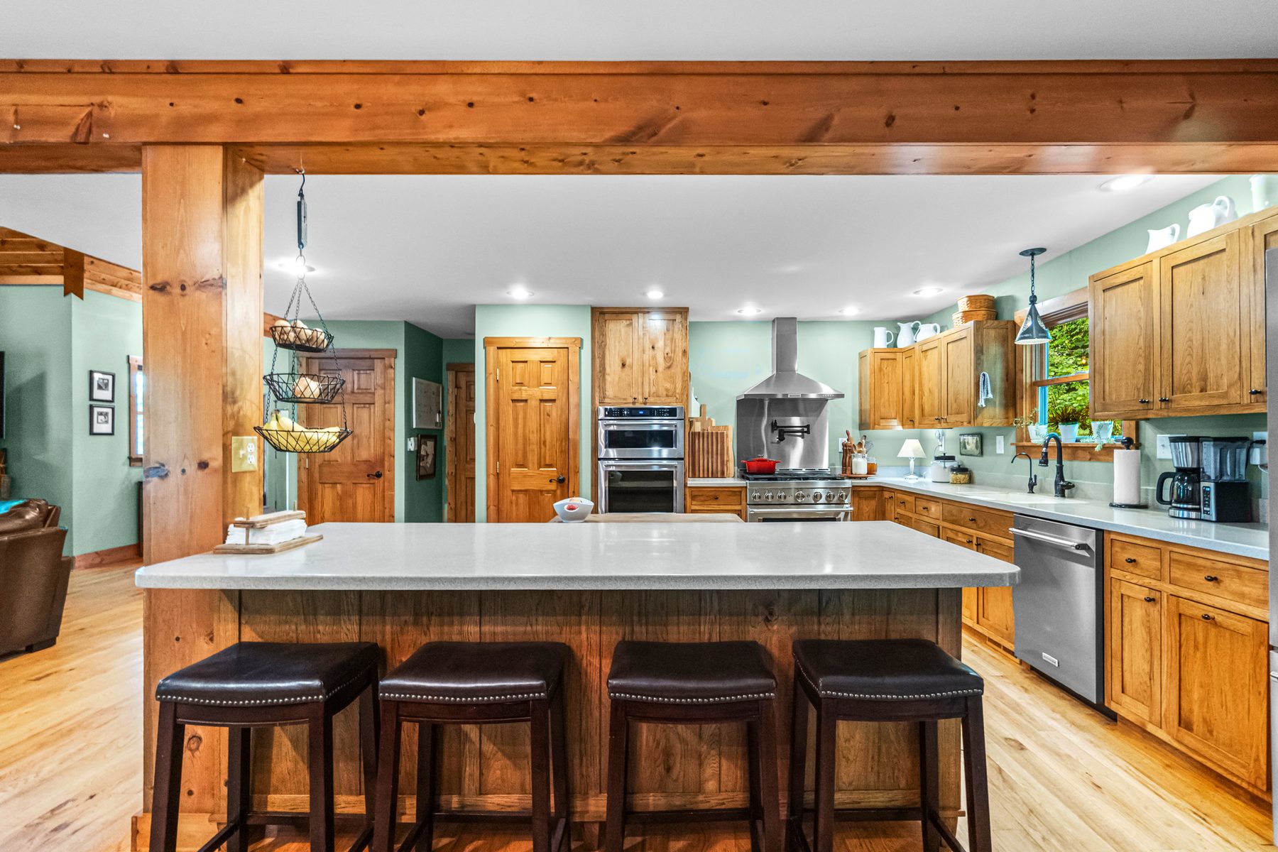 Kitchen island with four stools under exposed timber beams, with wall ovens and cabinetry beyond