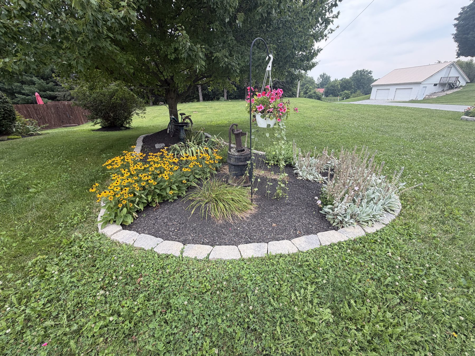 Landscaped island bed in the lawn with a decorative hand pump, yellow flowers and a stone border