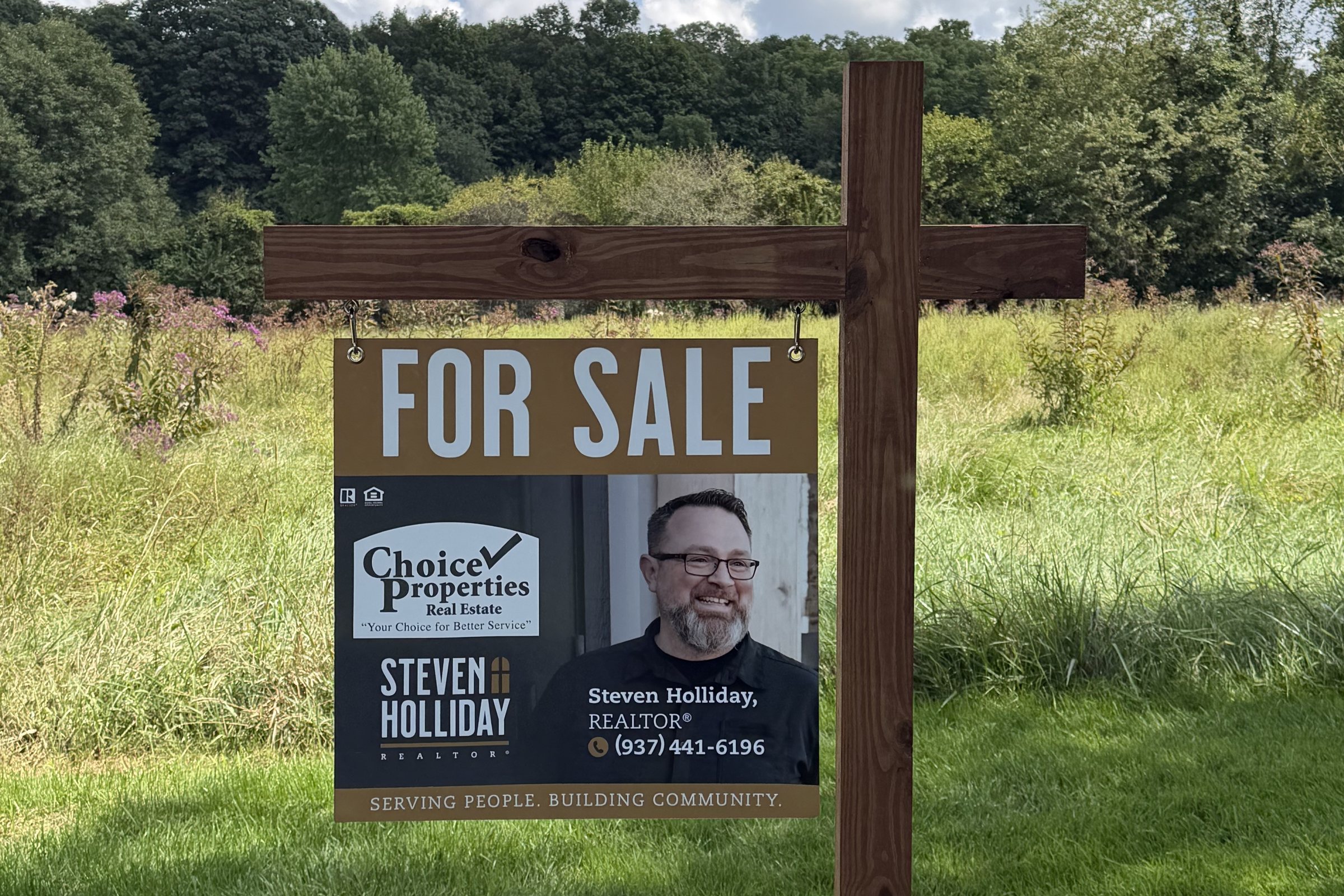 A Steven Holliday, REALTOR® with Choice Properties Real Estate For Sale sign in a mown field with a treeline behind it.
